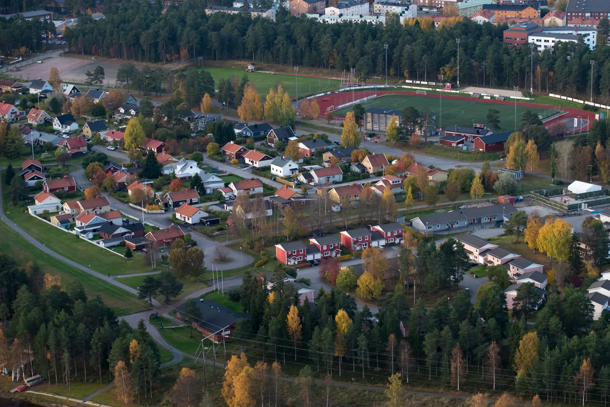 Flygbild över ett villaområde med trädgårdar och höstfärgade träd, med en idrottsplats i bakgrunden.