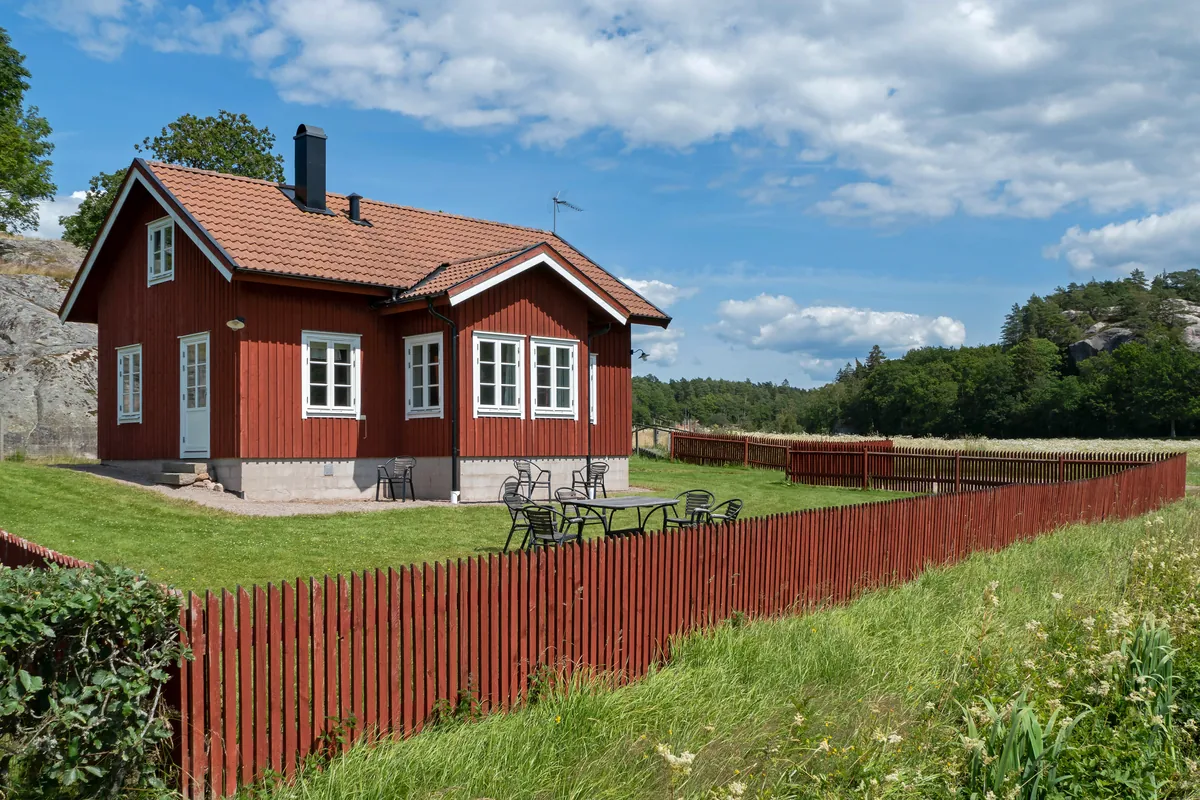 Rött trähus med vita knutar och rött staket på en grön gräsmatta, skog i bakgrunden.
